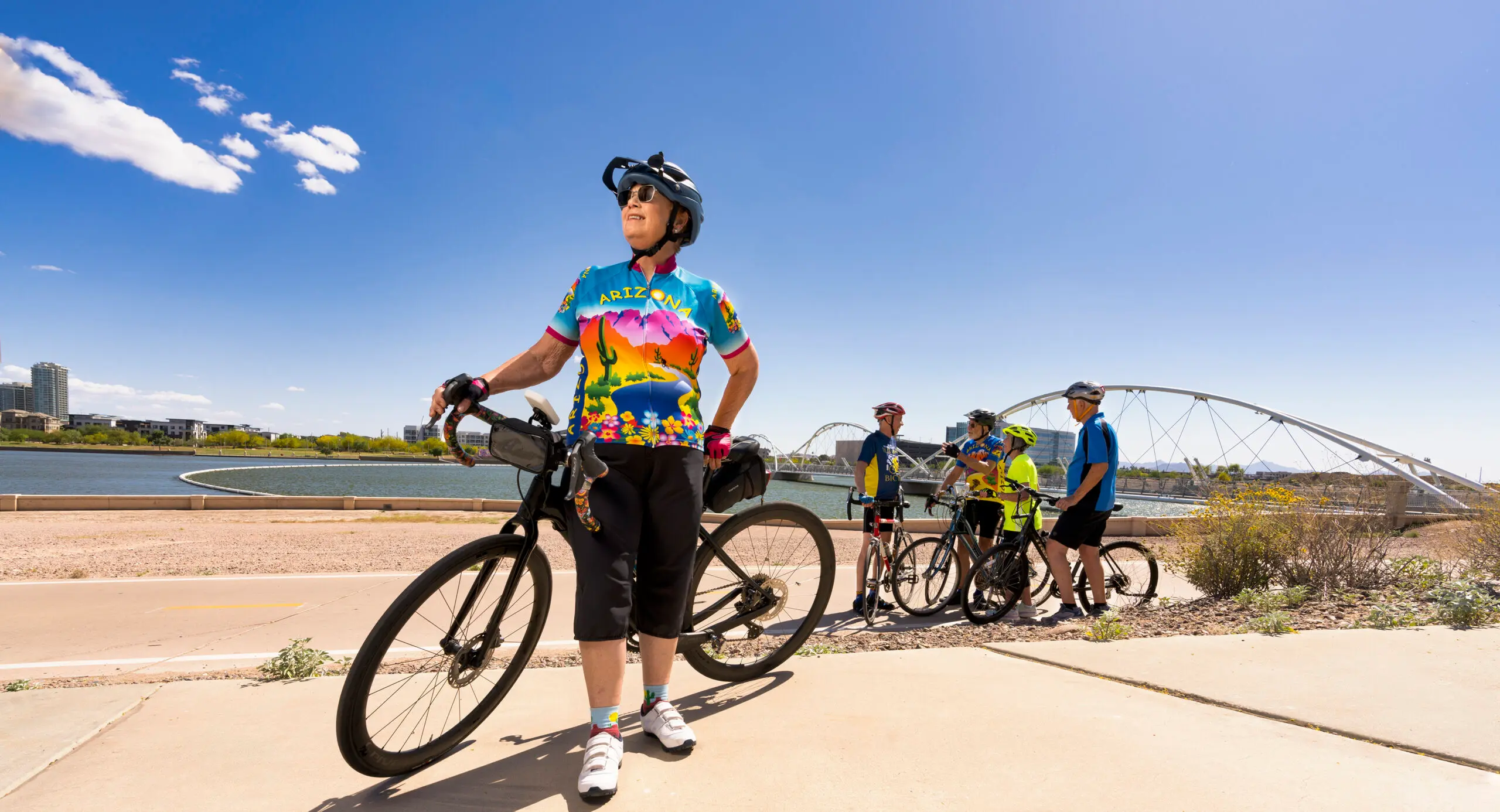 An older person stands with a bike in the foreground of the photo. A group of bikers stands in front of a picturesque lake in the background.